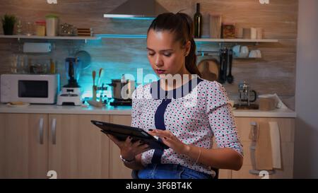 Thoughtful freelance woman using tablet pc while working overtime in home kitchen. Busy focused employee using modern technology network wireless doing overwork for job reading writing, searching Stock Photo