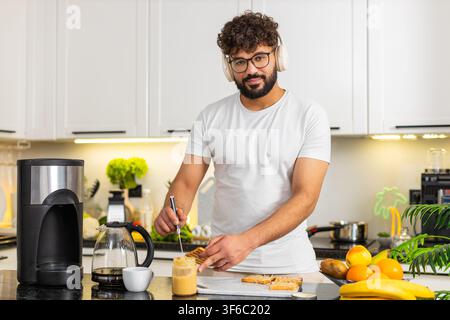 Arabian man dances with headphones in kitchen spreading peanut butter on toast for breakfast. Young guy feels happy preparing quick morning meal with coffee aroma and a tasty healthy routine at home. Stock Photo