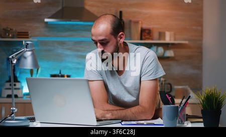 Entrepreneur sitting in remotely office working on laptop computer holding head with hands looking down. Busy focused employee using modern technology network wireless doing overtime for job reading Stock Photo