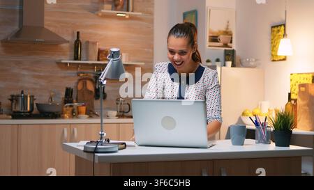 While working from home, the woman reacts with delight after reading excellent news on her laptop while working during an overtime session. Stock Photo