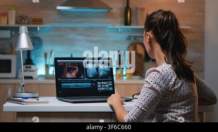 Woman edits video footage and audio on a professional laptop while completing a creative media project from her kitchen late at night. Stock Photo