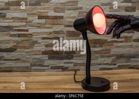 Black robotic or Prosthesis hand reaches toward a glowing yellow bulb in a modern desk lamp against a stone wall, useful for concepts of AI, innovatio Stock Photo