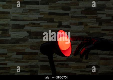 Black robotic or Prosthesis hand reaches toward a glowing yellow bulb in a modern desk lamp against a stone wall, useful for concepts of AI, innovatio Stock Photo