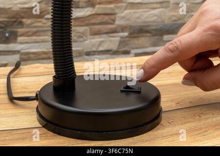 Hand touches switch on black desk lamp. Lamp base sits on wooden surface. Stone wall background adds rustic feel. Ideal for workspace or study room vi Stock Photo