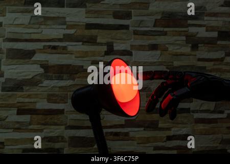 Black robotic or Prosthesis hand reaches toward a glowing yellow bulb in a modern desk lamp against a stone wall, useful for concepts of AI, innovatio Stock Photo