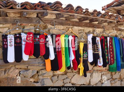 Traditional socks in various colors and patterns are hung on metal hangers against a rustic stone wall, featuring wooden beams and a sunny atmosphere Stock Photo