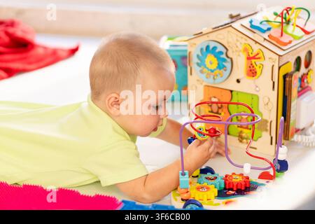 Baby boy lies on his stomach and explores a colorful wooden activity cube with bead mazes, gears, and movable pieces during tummy time in a bright hom Stock Photo