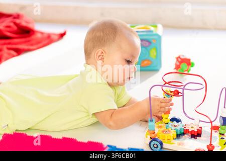 Infant boy lies on his stomach and carefully moves colorful beads along a wooden wire maze, practicing coordination and focused play during tummy time Stock Photo