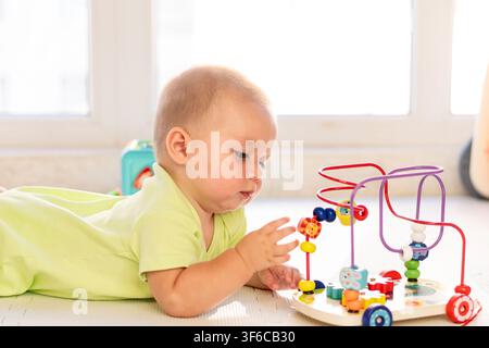 A baby boy reaches toward colorful beads on a wooden maze toy while lying on his stomach, exploring movement and coordination during bright indoor tum Stock Photo