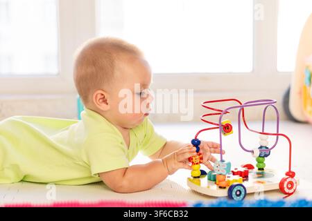 An infant boy uses both hands to move beads on a colorful wooden maze toy while lying on his stomach, building fine motor skills during playtime in a Stock Photo