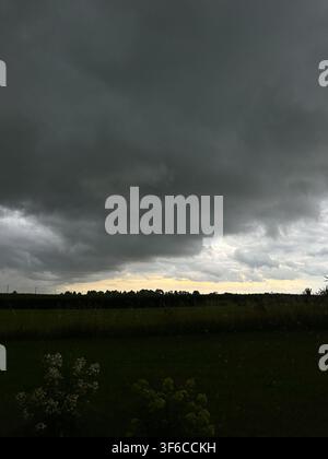 Dramatic dark storm clouds gather over a peaceful rural landscape moments before heavy rain. Atmospheric weather scene with open fields, moody sky, an Stock Photo