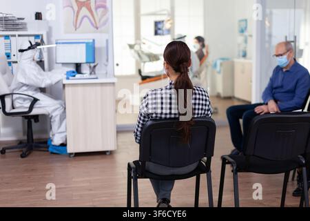 As staff coordinate care, the dentist speaks with a female patient in the clinic corridor while protective clothing is worn and other dental appointments continue around the reception area. Stock Photo