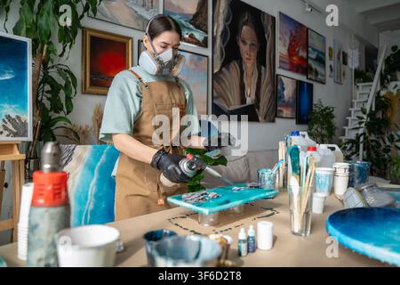 Focused craftswoman creating fluid art painting in gallery workshop using gas torch and epoxy resin. Stock Photo