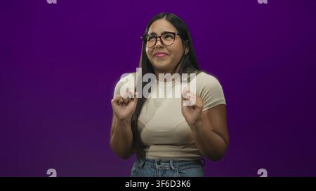 Young woman with glasses and hands raised, recoiling with a disgusted facial grimace, wearing beige tee and jeans in a purple studio; disgust avoidanc Stock Photo