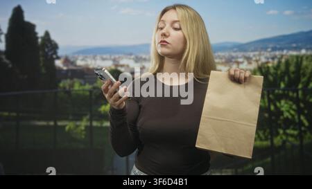 Woman holding smartphone and brown paper delivery bag, checking phone screen with a puzzled expression while standing by railing overlooking city on s Stock Photo