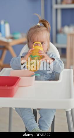 Toddler girl drinking from yellow sippy cup, holds cup to mouth in kitchen highchair with bowls and spoon on tray; calm domestic morning. Stock Photo