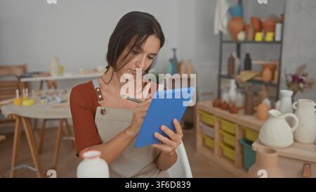 Woman holds blue tablet and taps with stylus in pottery studio, hands visible and focused on screen while surrounded by shelves of ceramic vases; crea Stock Photo