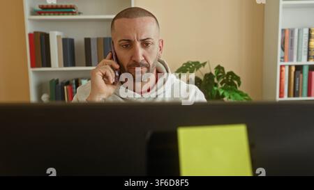 Man holding smartphone to ear at desk in building, glancing at computer screen, typing on keyboard and frowning; concern productivity. Stock Photo