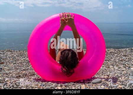 Beach ring summer woman relaxing with a bright pink inflatable float on a pebble beach overlooking the calm blue sea Stock Photo