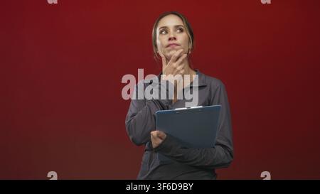 Woman holding a blue clipboard touches her chin in studio with red backdrop and casual sports top; thoughtful reflection. Stock Photo