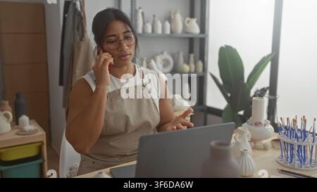 Woman talking on phone, gesturing with hand while using laptop, surrounded by ceramic vases and brushes in pottery studio; concentration creativity. Stock Photo