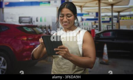Woman wearing apron tapping a tablet at a gas station pump, smiling and checking orders on a handheld device; calm focus service. Stock Photo