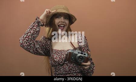 Young woman holding vintage camera with leather case, waving right hand and smiling while wearing straw hat and floral dress in studio; joyful spontan Stock Photo