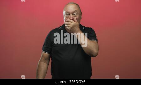 Hispanic senior man covers mouth with hand, clenches fist and looks startled in red studio; surprise silence. Stock Photo