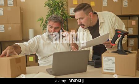 Two men with hand on parcel, holding phone to ear and leaning over laptop in building; teamwork productivity. Stock Photo