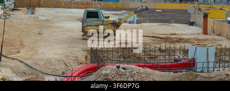 Workers are preparing the site for new building construction. A small machine is moving earth while pipes are shown on the ground. The project is loca Stock Photo