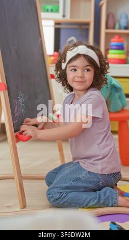 Girl kneeling drawing on a chalkboard with hands on chalk in a studio playroom, wearing headband and jeans while smiling at toys; playful curiosity. Stock Photo