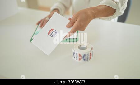 Woman hand offering ballot envelope beside roll of voted stickers and clipboard on table at building polling station; pride civic duty. Stock Photo