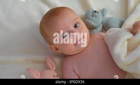 Baby girl lying on side holding soft blanket and knitted toy on white bed in studio; gentle bonding calm. Stock Photo