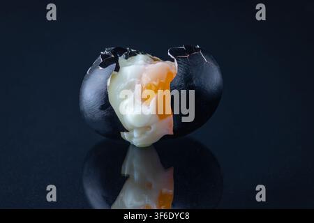 Horizontal dramatic studio still life of two cracked black painted eggs with orange yolk visible, reflected on a dark glossy surface. The paired crack Stock Photo