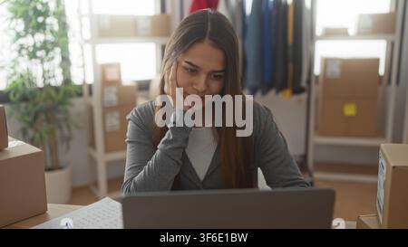 Woman at laptop with hand on cheek checking shipping boxes and orders in studio; thoughtful planning. Stock Photo