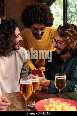 Group of man friends in a bar having fun, drinking beer, using one phone to watch social media. Stock Photo