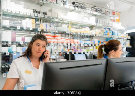 Female pharmacist assists a customer by telephone at a pharmacy counter, with prescription medicine boxes on shelves and a colleague working at comput Stock Photo