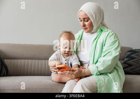 Muslim mother wearing a hijab sits on a sofa with her baby, helping the child explore a smartphone in a comfortable family home setting. Stock Photo