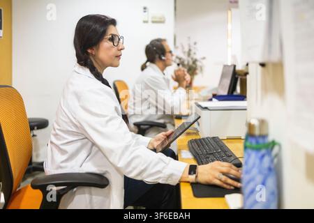 Female doctor in a white coat uses a computer and tablet at a medical clinic reception desk while a male colleague works in the background. Stock Photo