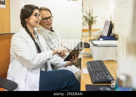 Female and male doctors in white coats review patient information on a tablet at a desk in a medical clinic, using digital technology for healthcare c Stock Photo