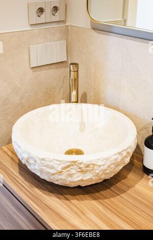 Close-up of a modern apartment bathroom featuring a textured stone vessel sink, tall brass faucet, wood vanity, wall mirror, and electrical outlet in Stock Photo