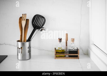 Modern apartment kitchen interior with wooden and black cooking utensils in a metal holder beside olive oil and vinegar bottles on a white marble coun Stock Photo