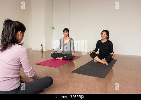 Three Japanese women sit cross-legged on yoga mats during a calm indoor yoga class, practicing meditation and mindful breathing in a bright minimalist Stock Photo
