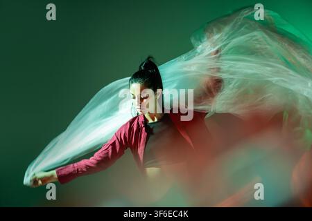Latin woman dancer performs an expressive movement with translucent flowing fabric, illuminated by green and red light in an atmospheric studio portra Stock Photo