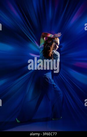 Young woman poses in a dance movement with raised arm, surrounded by colorful blue and purple abstract light rays in a dark studio, creating an energe Stock Photo