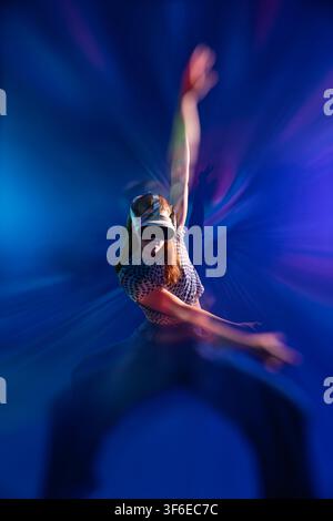 Young woman wearing a virtual reality headset performs an expressive dance pose with raised arms, surrounded by vivid blue and purple light trails and Stock Photo
