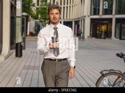 Young businessman in a white shirt and tie uses a smartphone while walking through a modern business district, surrounded by office buildings and an u Stock Photo