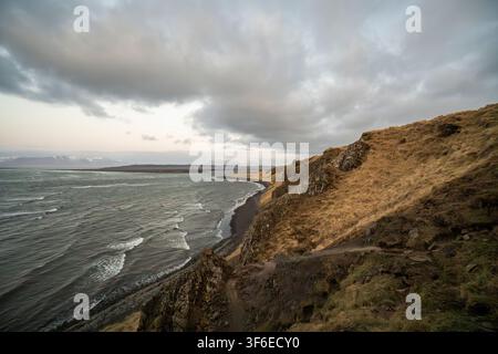 Dramatic view of rugged coastal cliffs above a black sand beach in Iceland, with waves rolling across the North Atlantic beneath heavy storm clouds du Stock Photo
