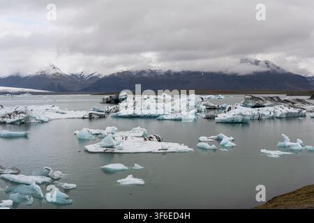 Floating icebergs drift across a glacial lagoon beneath snow-covered mountains and dramatic gray clouds in Iceland, creating a cold and tranquil Arcti Stock Photo