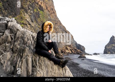 Traveler wearing a fur hooded winter coat sits on rugged coastal rocks beside a black sand beach in Iceland, surrounded by dramatic cliffs, sea stacks Stock Photo